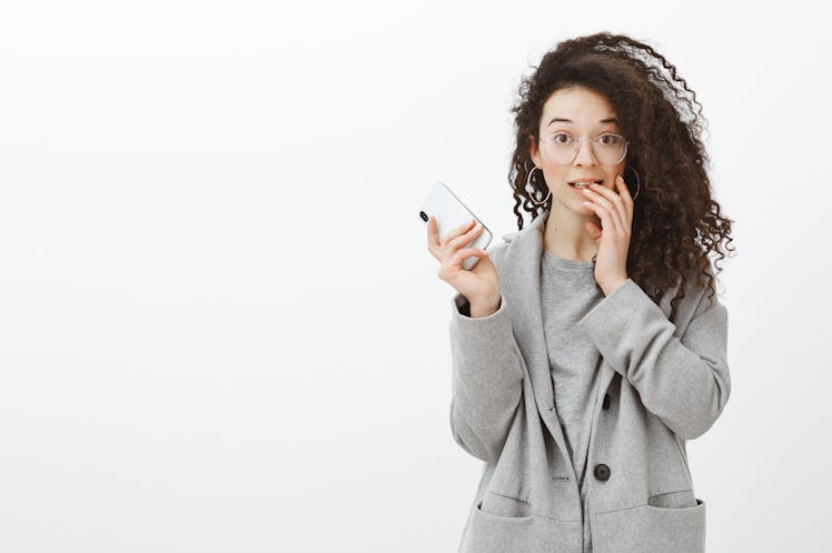 Portrait of embarrassed cute curly-haired female student in stylish grey coat and glasses, holding s...