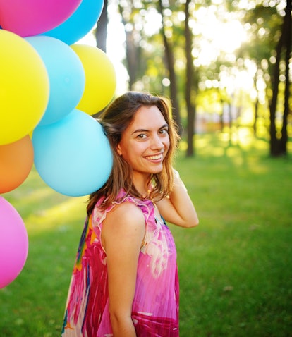 Beautiful, cheerful young woman in a bright dress with colorful balloons in a park with green grass ...