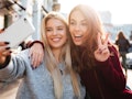 Two joyful cheerful girls taking a selfie while sitting together at cafe and showing peace gesture o...