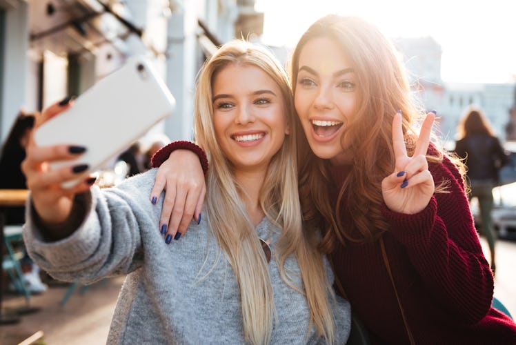 Two joyful cheerful girls taking a selfie while sitting together at cafe and showing peace gesture o...