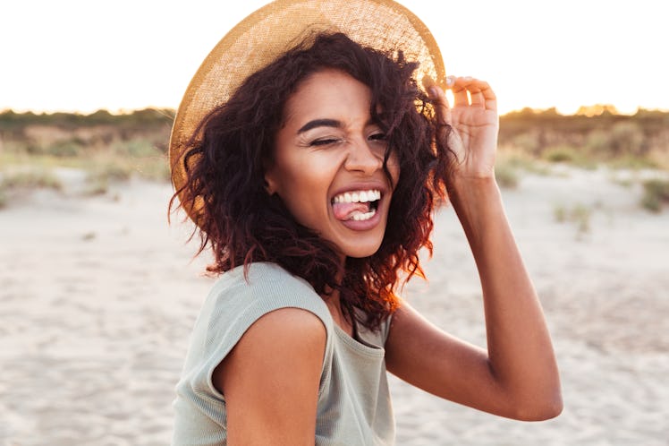 Close up of cheerful young african girl in summer hat looking at camera and winking at the beach