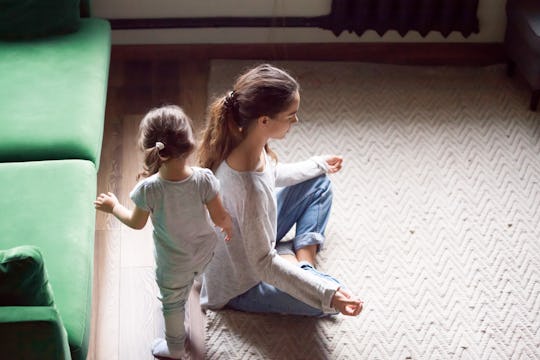 Young single mother doing yoga exercise, meditating while little preschool daughter running, playing...