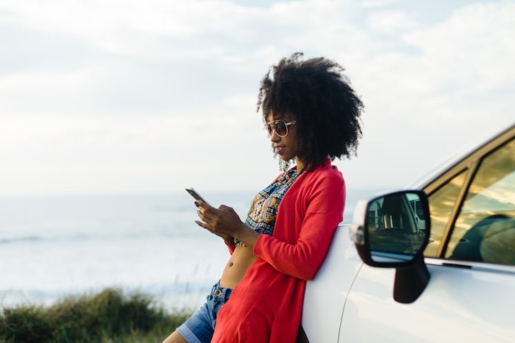 Fashionable afro hair woman on vacation texting on smartphone towards the sea. Stylish black model r...