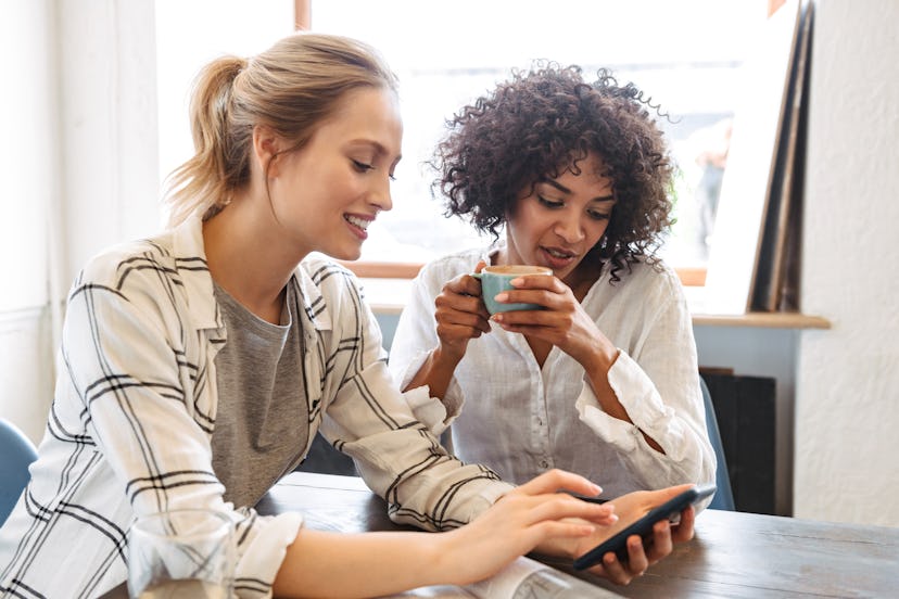 Two happy young women friends sitting at the cafe, having coffee, using mobile phone