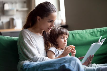 Young mother teaching daughter holding book sitting on sofa, mom or babysitter embracing little girl...