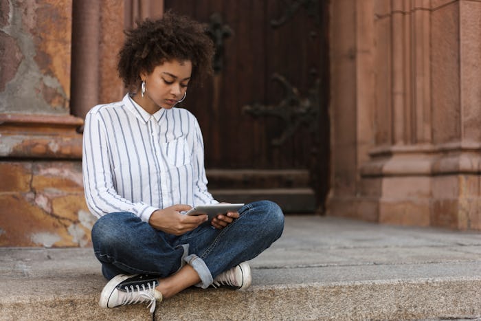 African-American girl sitting on the steps near the building. She works on a tablet.