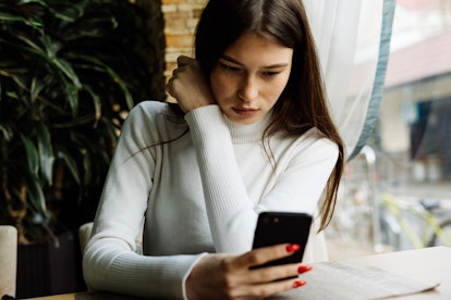 beautiful young dark-haired girl in a white sweater sits in a cafe and looks in a smartphone