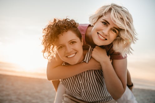 Portrait of a smiling young woman giving her girlfriend a piggyback while having fun together at a b...