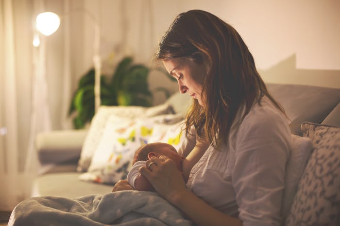 Young beautiful mother, breastfeeding her newborn baby boy at night, dim light. Mom breastfeeding in...