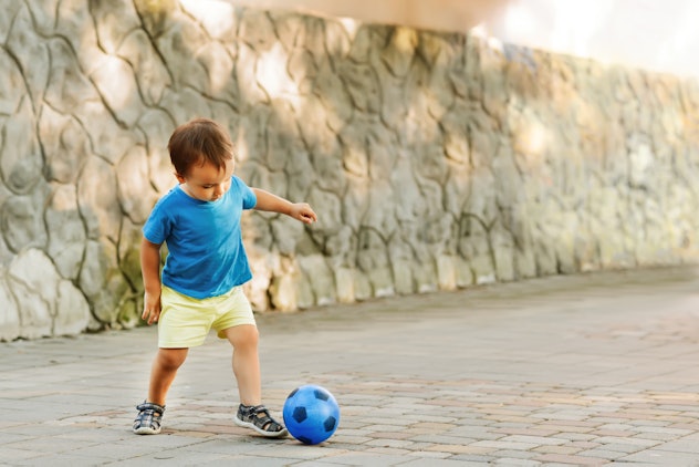 Little boy in blue shirt and yellow shorts playing football with blue tiny toy soccer ball in a sunn...