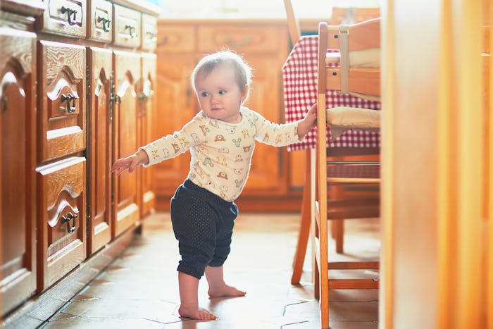 Baby girl standing on the floor in the kitchen and holding on to furniture. Little child oulling up ...
