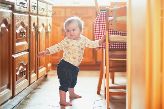 Baby girl standing on the floor in the kitchen and holding on to furniture. Little child oulling up ...