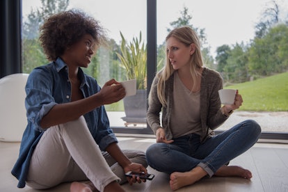 Two young laughing multiethnic women sit on the floor near the window and enjoy while drinking coffe...