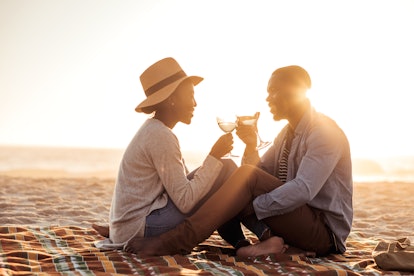 Romantic young African couple toasting each other with wine while sitting together on a sandy beach ...