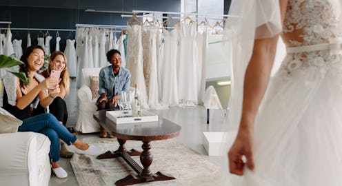 Women taking photographs of a female friend trying on wedding dress. Women in wedding dress fitting ...