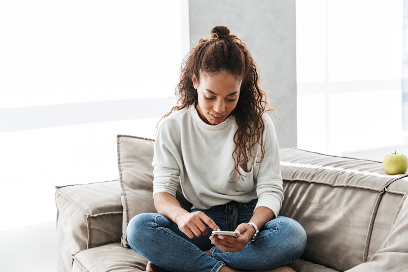 Photo of adorable african american woman using cell phone while sitting on couch in bright apartment