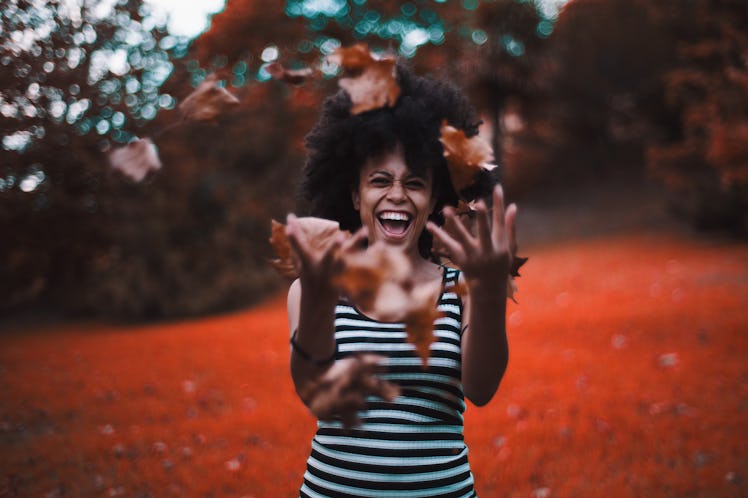 Curly laughing African-American girl in a striped dress is fooling and throwing up a heap of dry aut...