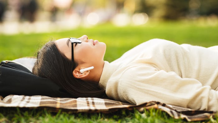 Girl listening music in airpods with closed eyes, lying on blanket in park