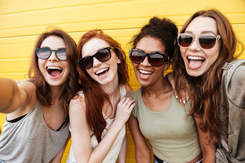 Image of young happy women friends standing over yellow wall. Looking at camera make selfie.