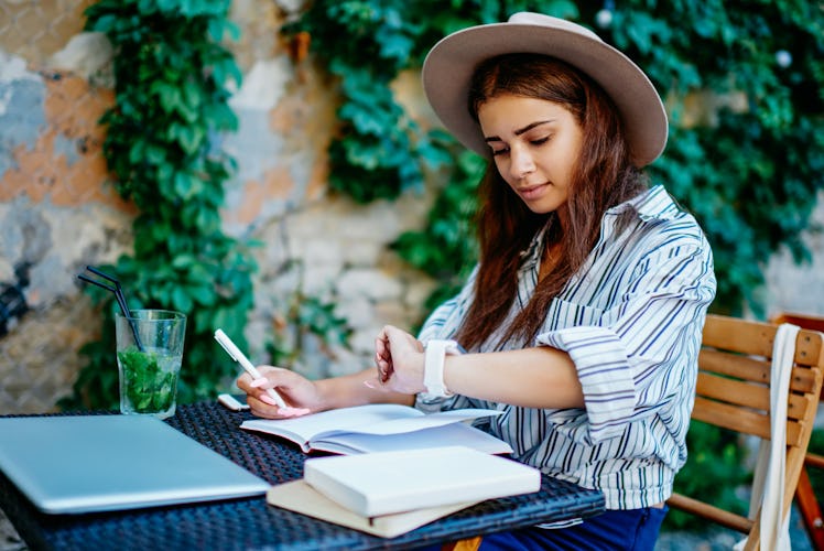 Pensive female blogger writing in notebook checking notification on modern designed wearable compute...