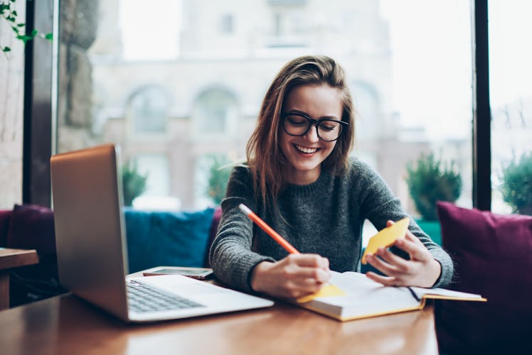 Positive female student in trendy eyeglasses enjoying time with modern notebook at coworking space, ...