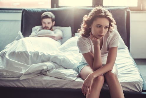 We have a problem. Young upset girl sitting on the edge of the bed, against her boyfriend, lying in ...