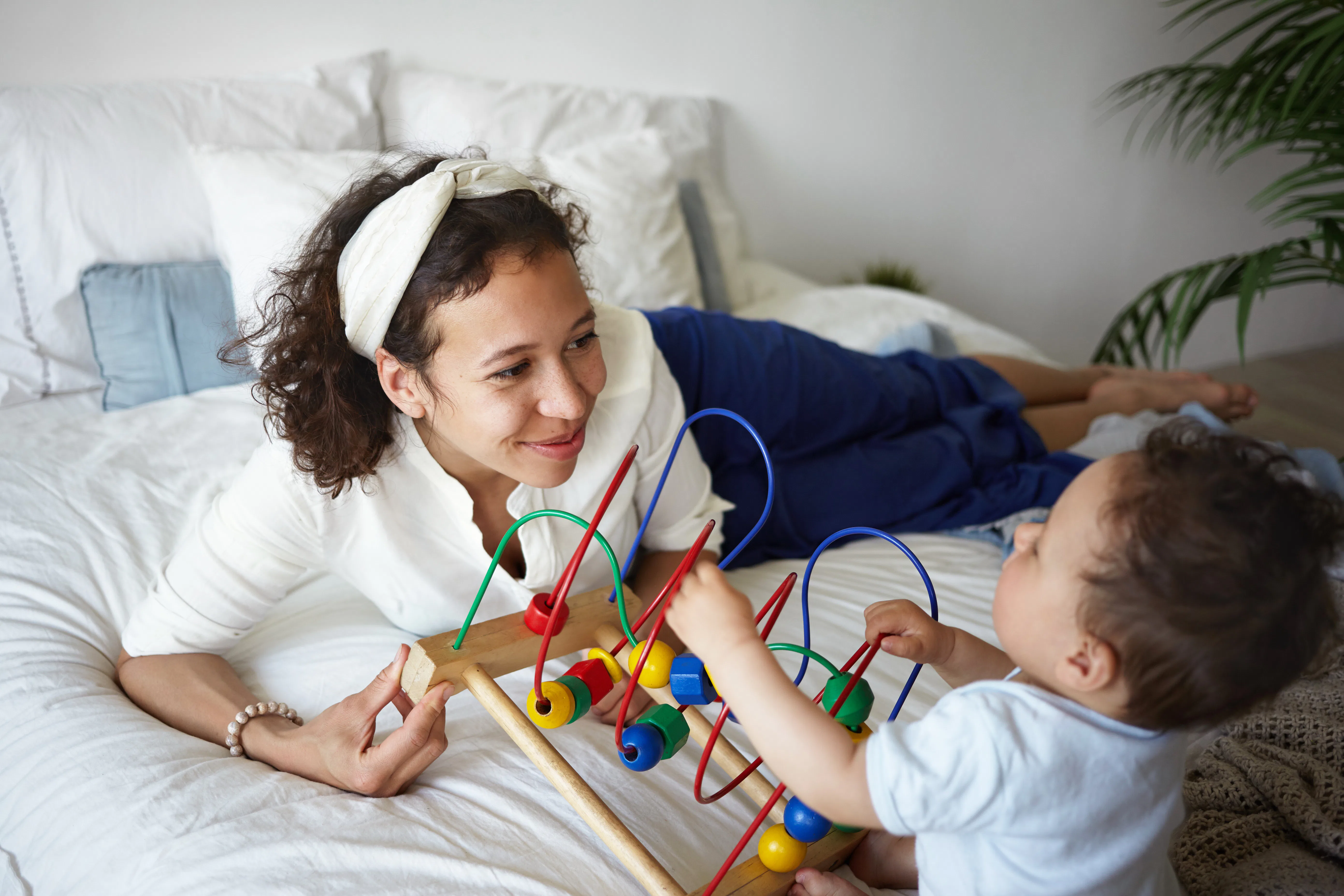 Picture of stylish young Hispanic female smiling joyfully, teaching her baby infant how to play new ...