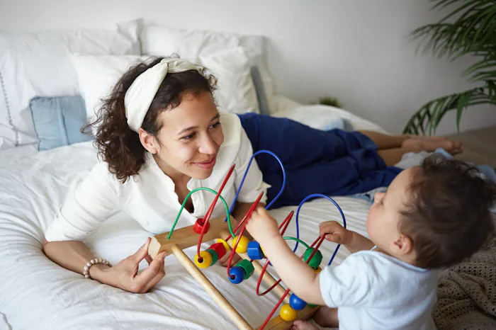 Picture of stylish young Hispanic female smiling joyfully, teaching her baby infant how to play new ...