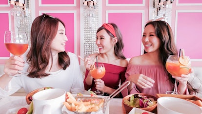 Three friends laugh and hold their cocktails at a pink brunch spot on a sunny morning.