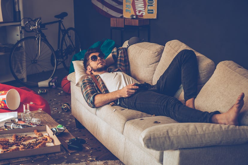 A man lying on his couch with sunglasses while there is a huge mess around him
