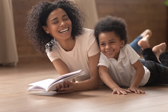 Overjoyed young black mother have fun reading book with cute little son laughing lying on floor toge...