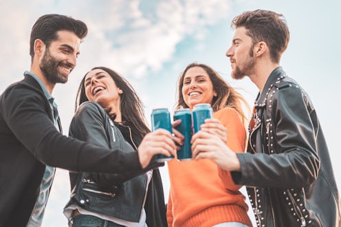 Two guys and two girls smiling and toasting with canned beers outdoor. Carefree youth having fun out...