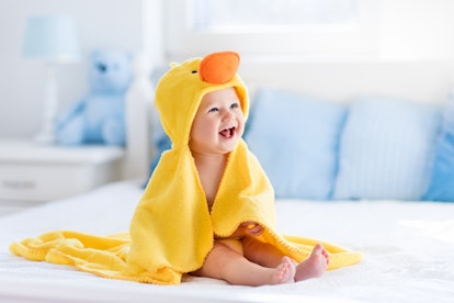 Happy laughing baby wearing yellow hooded duck towel sitting on parents bed after bath or shower. Cl...