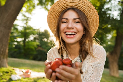 Close up of happy young girl in summer hat spending time at the park, holding heap of strawberries