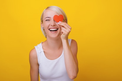 Woman in love, holding a littlle paper heart over eye, standing over yellow background 