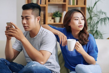 Beautiful Asian woman with coffee leaning on boyfriend absorbed with smartphone and looking bored