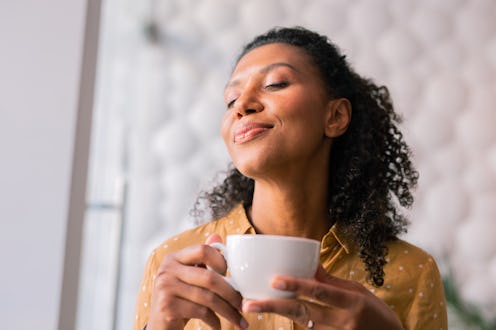 Feeling joyful. Curly dark-haired appealing woman wearing yellow blouse feeling joyful while drinkin...