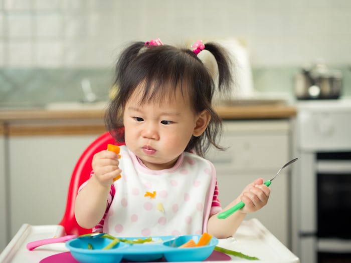 Toddler with pigtails eating peas and carrots at home.