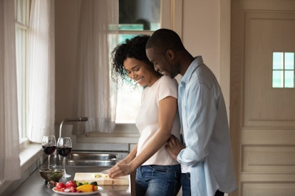 A happy, trendy couple embraces while chopping vegetables in their kitchen on a sunny day.