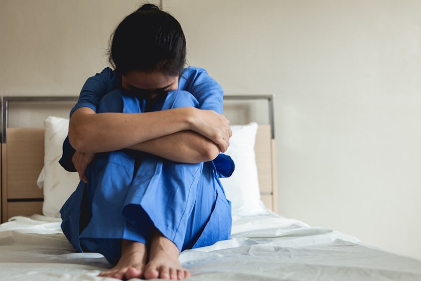 Woman in scrubs sits with head tucked into knees on bed.