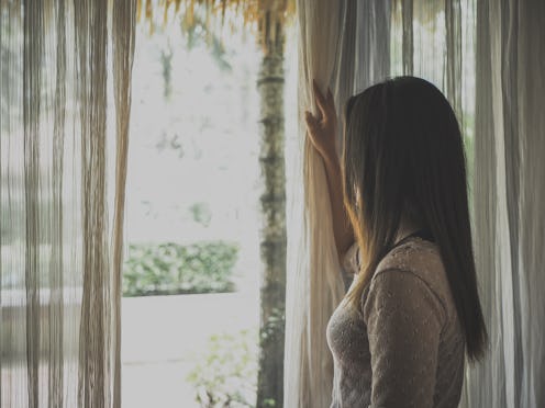 Sad woman holding the curtains open to look out of a large light window at home. Positive and aspir...