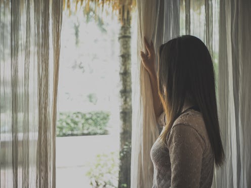 Sad woman holding the curtains open to look out of a large light window at home.  Positive and aspir...