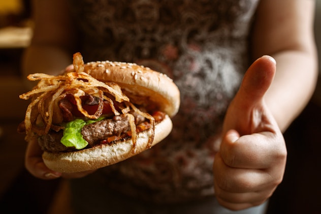 Unrecognizable pregnant woman enjoy her burger. Girl holding taking onion hamburger in one hand and ...