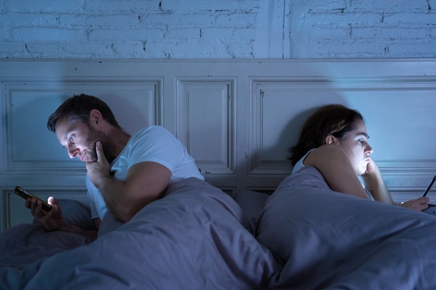 Young couple in bed late at night using smart phones obsessed with games, social media and apps igno...