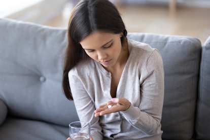 Pensive girl sit on couch holding medicines and glass of water thinking of taking medications, depre...