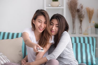 A queer couple sitting on the couch, with a remote in hand, watching movies on the TV. 