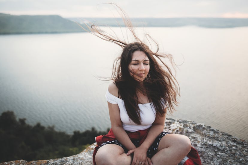Travel and Meditation. Hipster girl with windy hair sitting and meditating on top of rock mountain w...