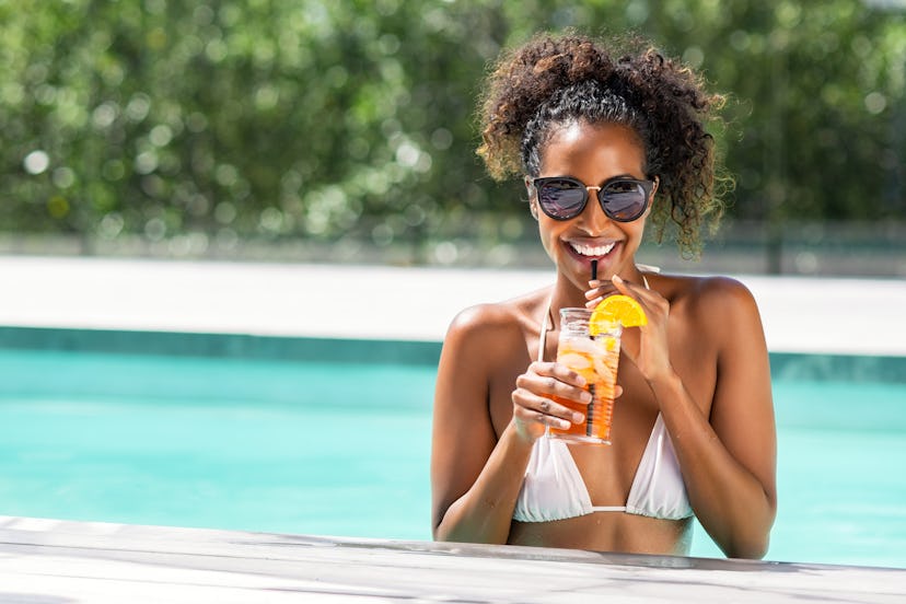 Portrait of happy fashion woman with sunglasses standing in swimming pool drinking cocktail. Beautif...