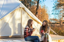 Two happy friends drinking coffee while relaxing near canvas bell tent in the morning in a forest