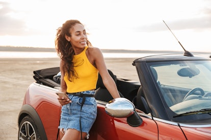 Picture of a smiling happy optimistic young pretty woman standing near car at the beach using mobile...
